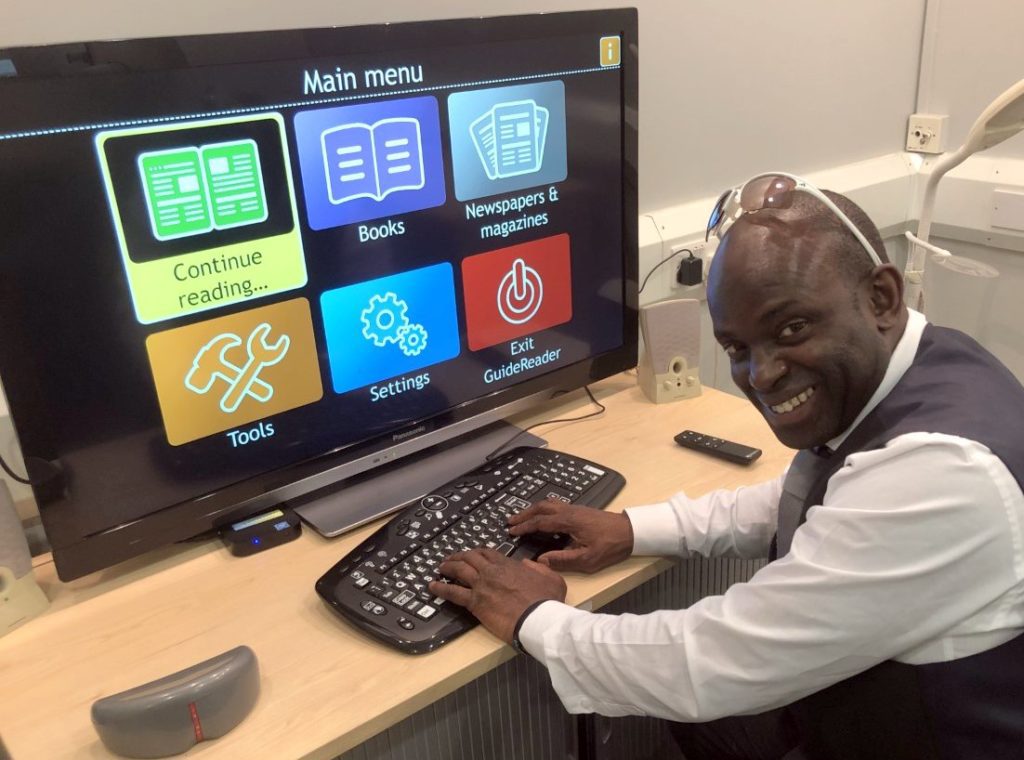 Man with sight loss at workstation with large display screen showing large menu icons. He's smiling as the tech makes his his working life easier.